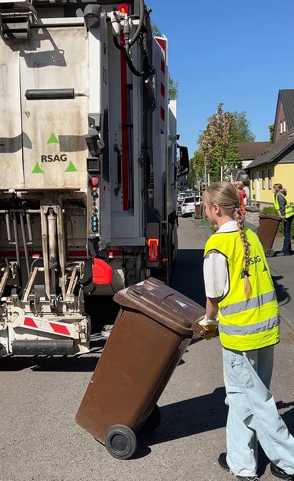 Ein Mädchen schiebt eine braune Biotonne zu einem Müllfahrzeug. 
