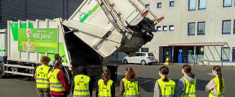 Acht Mädchen stehen vor einem Müllwagen und beobachten, wie dieser sich öffnet. 