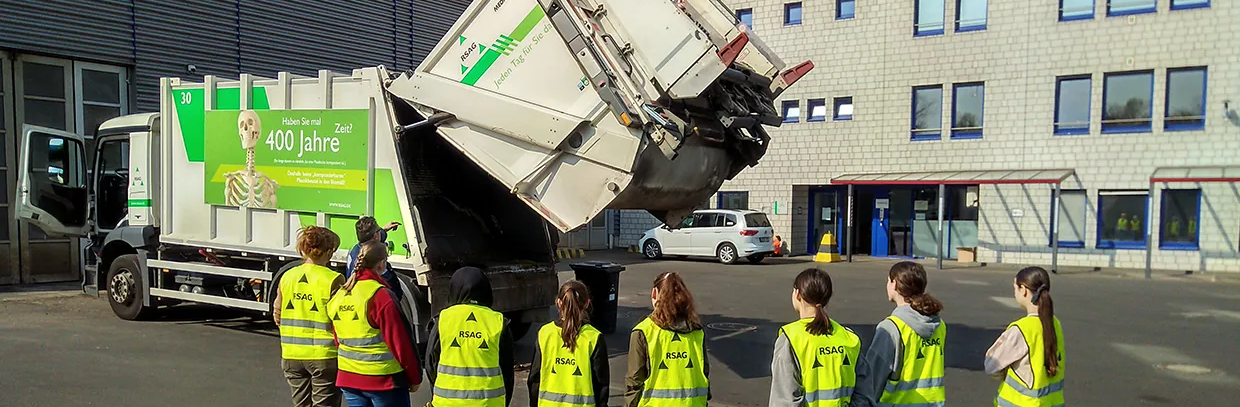 Acht Mädchen stehen vor einem Müllwagen und beobachten, wie dieser sich öffnet. 