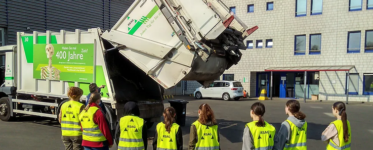 Acht Mädchen stehen vor einem Müllwagen und beobachten, wie dieser sich öffnet. 